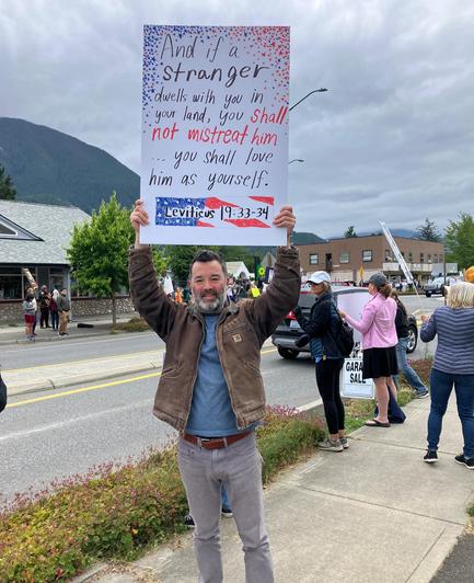 Photo of a demonstrator at a small town demonstration (No Kings?) holding a board above his head and smiling 

Board reads - 
And if a stranger dwells with you in your land, You Shall Not Mistreat Him.
…You Shall love him as yourself.
Leviticus 19.33.34