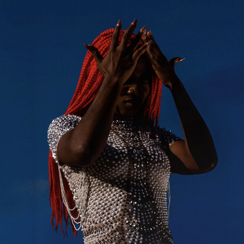 Aunty Rayzor wearing a very bejeweled top, with red braids, in front of a blue background... hands up and on her forehead, fingers wide.