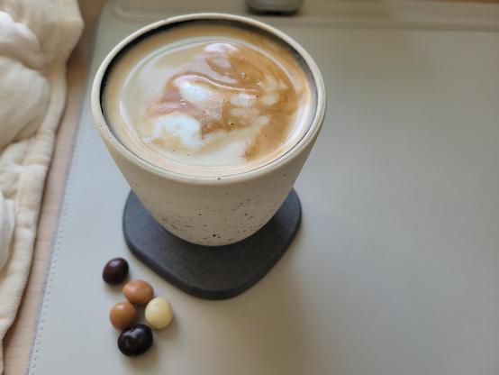 12-ounce, handle-less, off-white ceramic cup with a pumpkin-spice coffee latte, on a gray diatomaceous earth coaster on a white desk mat, on a maple desk. The clay of the cup has many tiny flecks of black and blue. The interior of the cup has a dark-colored glaze. To the bottom-left of the cup are five chocolate-covered espresso beans, in light-brown, medium-brown, and dark-brown colors.