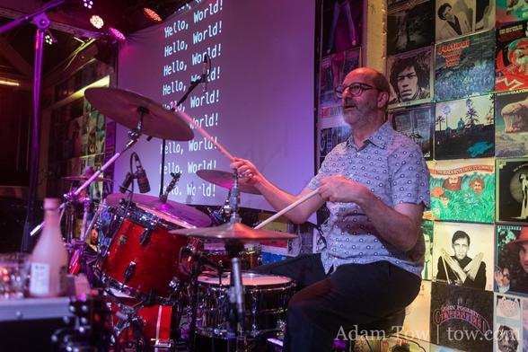 John Fox playing drums with “Hello, World!” slide in the background. Photo by Adam Tow.