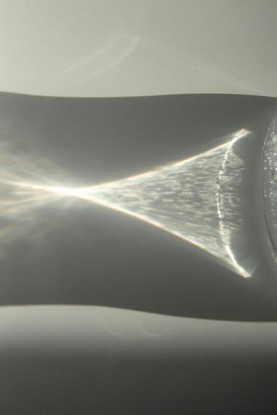 Gebündelter Lichtstrahl, der durch ein Wasserglas auf eine weiße Oberfläche fällt – die Lichtfokussierung erinnert an die Form eines Laserstrahls. Foto von Jill Burrow via Pexels.