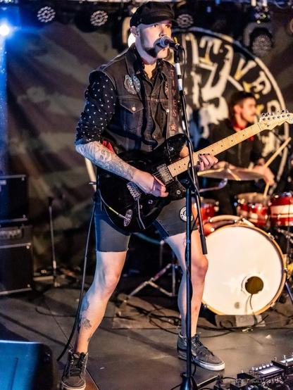 A musician performs on stage, playing an electric guitar and singing into a microphone. He wears a black vest adorned with patches and shorts, and has visible tattoos on his arms. A drummer is partially visible in the background, with drums set up behind him.