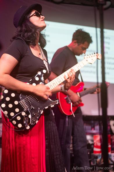 Shloka Kini wearing sunglasses and a black fedora, playing a black electric guitar with white polka dots. In the background Pavan Rajam playing electric guitar. Photo by Adam Tow.
