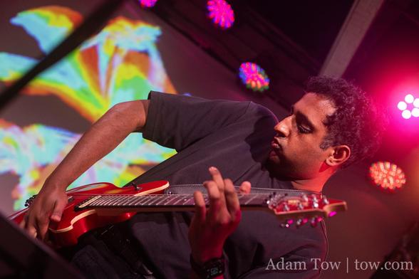 Pavan Rajan playing a red electric guitar. In the background multicolor abstract computer graphics projected on a screen. Photo by Adam Tow.