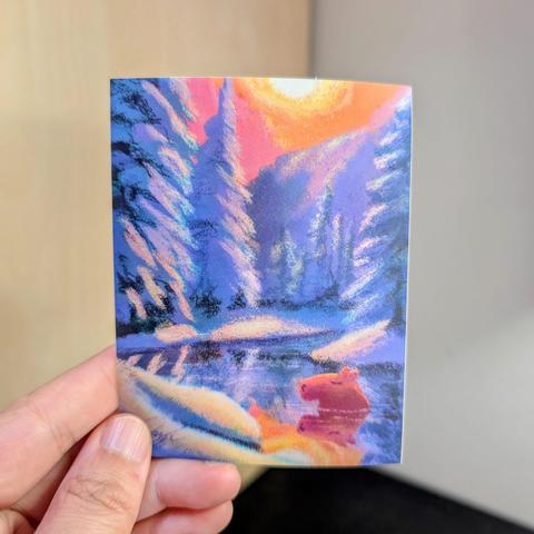 Photo of a left hand holding a rectangle sticker. The sticker is a matte painting of a capybara resting among snow and trees.