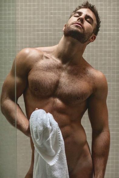 A muscular white man is slowly drying himself with a towel in the shower 