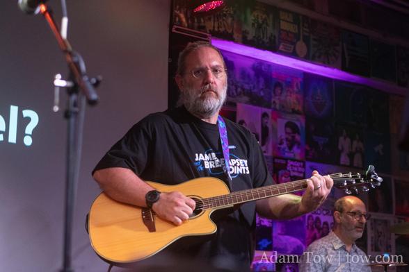 Josh Smith playing an acoustic guitar, wearing a James Dempsey and the Breakpoints t-shirt. John Fox is in the background. Photo by Adam Tow.