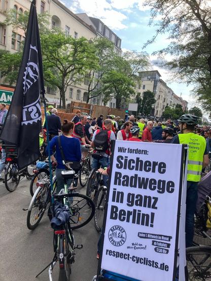 Demonstrierende mit Fahrrädern auf dem Savignyplatz. Im Vordergrund ist links ein Fahrrad mit Respect Cyclists Fahne zu sehen und rechts nimmt ein Anhänger mit Transparent ein Viertel des Bildes ein. Transparenttext: Wir fordern: Sichere Radwege in ganz Berlin!