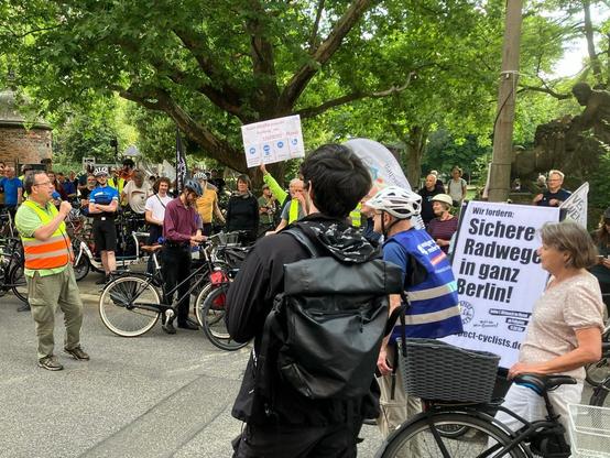 Eine Gruppe Radfahrender bei der Zwischenkundgebung auf dem Savignyplatz. Links im Bild ist ein Redner zu sehen. In der Bildmitte wird ein Plakat mit einer schematischen Straßendarstellung hochgehalten, auf dem zu lesen ist: Kantstraße braucht einen Radweg am sicheren Rand. Rechts zwischen weiteren Personen ist auch der Transparentanhänger zu sehen: Wir fordern: Sicher Radwege in ganz Berlin!