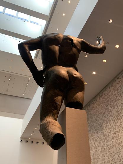 Reverse view of a bronze sculpture thought to depict Alexander of Macedon, aka Alexander the Great. The camera shot is from below looking up. The figure is nude, one arm resting on the hips, the other used to hold a spear or a sceptre.