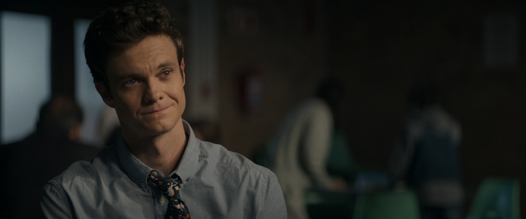 Cena do filme Novocaine. Ela mostra Nathan Caine (Jack Quaid) sentado em um ambiente interno. Nathan Caine está vestindo uma camisa azul clara e uma gravata com um padrão colorido. Ao fundo, há outras pessoas desfocadas, uma delas parece estar de pé e inclinada sobre uma mesa. O ambiente é iluminado de forma suave, criando uma atmosfera introspectiva.