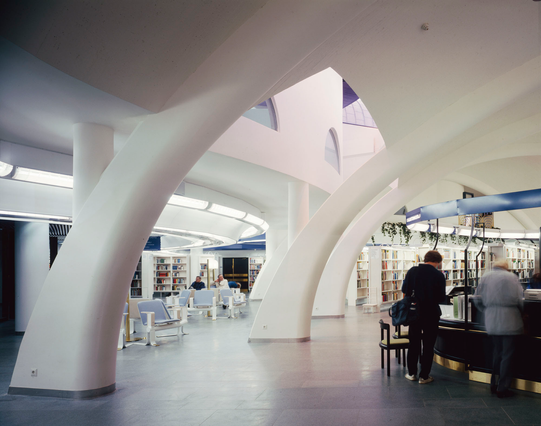 Es una foto del interior de la biblioteca Metso de Tampere. Se ven formas más bien redondas en columnas y son blancas, todo como en un único espacio