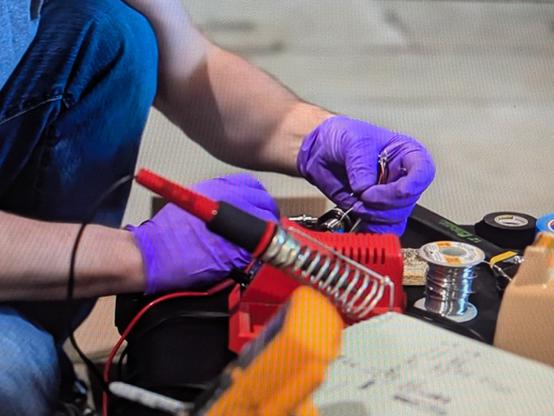 cheap red Weller soldering station. the knob on the front just adjusted the power level, not the temperature. someone is soldering while wearing purple gloves. off to the side is a spool of solder with a yellow color code indicating that it is leaded.