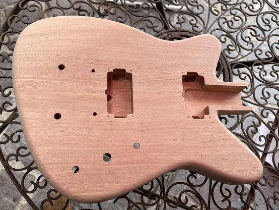 Der rohe, unlackierte Korpus (Body) einer E-Gitarre liegt auf einem Gartentisch aus schnörkeligem Eisen. Die Gitarre ist eine Reverse Offset und hat stark abgerundete Kanten.