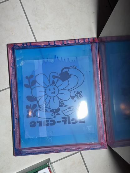A view of the printing frame, after applying a photosensitive emulsion, being prepared for exposing phase. A film positive of the illustration printed in black is placed on the screen and is exposed to a bright light source. The light hardens the emulsion except where the black ink of the design blocks it.