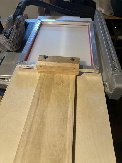 A screen printing setup featuring a wooden board positioned beneath a screen frame. The screen frame is secured with some screws that behave like a clamp. The setup is part of a screen printing press.