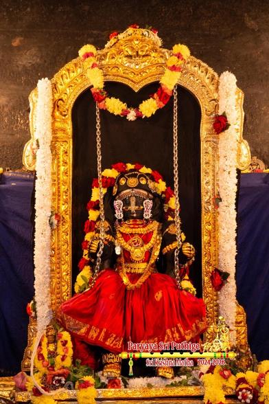 Idol of Hindu deity Krishna in red seated in a swing with both legs down and hanging in air above a gold platform; wearing gold shoes and lots of gold ornaments; hands seemingly holding the swing's chains (which hang down). A string of red and yellow flowers on Krishna's body going from his left waist to right waist up his body and around the back of his head.

A gold arch around (and slightly behind) Krishna from which the swing hangs down. Thick strings of white flowers hang down on both sides of the arch. A string of yellow and red flowers hangs in U shape at the top middle of the arch.

Blue cloth background.