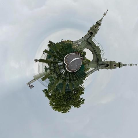 A panoramic view of a circular garden or park featuring a prominent archway with decorative towers. The scene is overcast, with greenery surrounding the area, and a circular tennis court visible in the center.