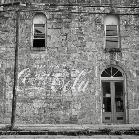 A black and white film photo. A weather-worn hand-painted sign that states “Drink Coca-Cola“ is seen on a very old stone building façade in Gonzales, Texas. Sunday, June 1, 2025.