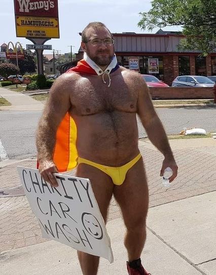 A man wearing only yellow briefs and a flag on his back is holding a sign saying "charity car wash"