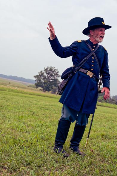 Man in blue blue coat and hat with white bear is talking, wearing a sword and boots standing on a lawn with a slanted horizon behind him.