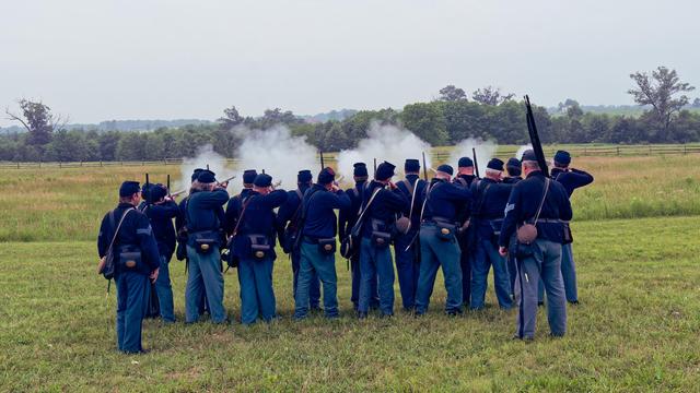 Soldiers with blue coats, pants and hats hold face away from the camera and hold black powder rifles up in the air as a cloud of white smoke rises in front of them.  They are standing on a close-cropped lawn with taller grass, a fence, a hedgerow and a line of distance fields past them.