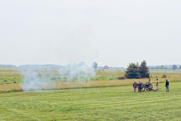 Five civil war reenactors surround a cannon on two wheels with a big cloud of smoke in front of them.  They look small compared to a landscape of lawn, fields, isolated trees, barns and such under a gray sky.