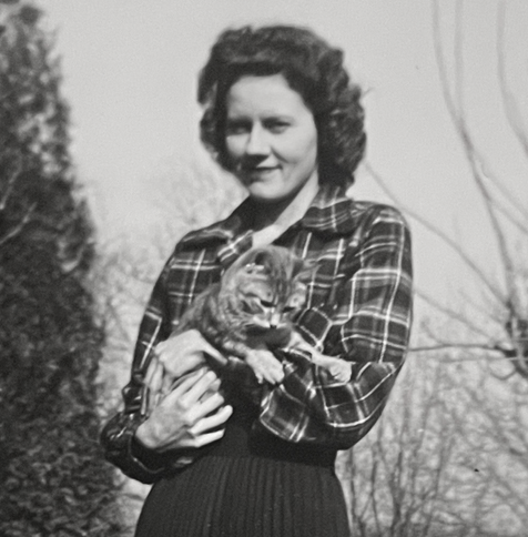 Black and white photo of a young light skinned woman wearing a plaid shirt and dark pleated skirt, holding a medium hair, tabby cat outside in the sun.