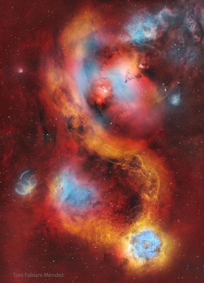 A deep, wide-field image of the Rosette Nebula region. The actual Rosette Nebula appears in the lower right, glowing in soft blue and white hues, with its central cavity carved out by bright blue stars from cluster NGC 2244. Delicate golden filaments stretch across the field, connecting other reddish nebulae in the frame. A large red nebula sits near the center but is not the Rosette. The background is filled with countless stars across the dark field of Monoceros.