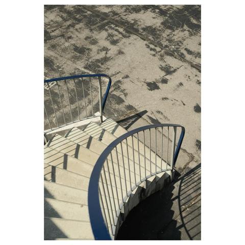 Block von oben herab auf eine geschwungene 60er Jahre Betontreppe. Weiße, vertikale Streben halten einen blauen Handlauf, der scharfe Schatten auf den fleckig geteerten Parkplatz wirft, auf dem die Treppe endet. 