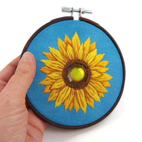Broderie d'une fleur de tournesol sur une toile bleue ciel tendue sur un cercle à broder en bois marron foncé.
Le cœur du tournesol est un morceau de CD fixé à la toile avec la technique de la broderie miroir, puis recouvert de résine époxy teintée jaune et marron.