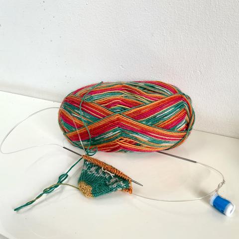 Angled view of a self-striping ball of sock yarn in yellow, orange, pink, and teal, as well as the start of a toe-up sock being knit magic loop on a long circular needle.