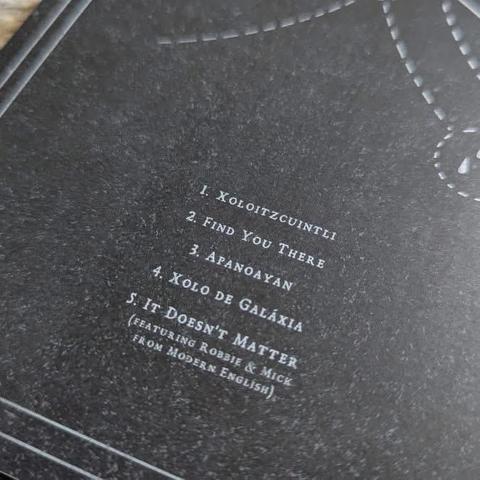 Eine Nahaufnahme einer Titelliste mit fünf Titeln, geschrieben in einer stilisierten Schriftart auf dunklem Hintergrund. Die aufgeführten Songs sind: 1. Xoloitzcuintli, 2. Find You There, etc.
