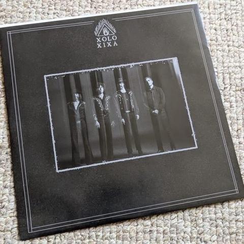 Ein Albumcover mit vier schwarz gekleideten Musikern, die in einer Reihe stehen. Der Hintergrund ist dunkel, und das Gesamtdesign hat eine Vintage-Ästhetik. Im oberen Bereich steht der Titel "XOLO XIXA".