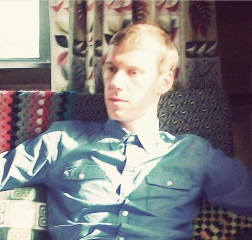 This is a picture of Cheapskate in a blue shirt sits on a patterned chair, dappled sunlight on their face, with floral curtains in the background. Calm atmosphere.