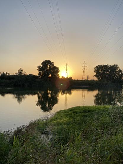 Foto del Río al atardecer, tomada hacia las 21:30. Se ve un rayo de sol en medio de la foto y una especie de pequeña isla de hierba en la orilla más próxima. En la orilla del otro lado, la silueta oscura de los árboles y postes de luz a lo lejos 