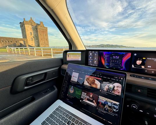 A laptop open in a car with a small screen in the dash showing the standard Mac System Settings. Outside we see the mouth of the River Tay, and Broughty Ferry castle, a squarish, boxy building.