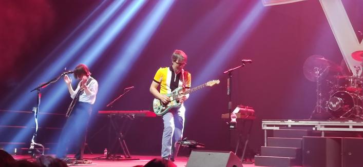 Rivers Cuomo alongside of the band playing guitar on stage. Brian on the guitar to the left and Patrick on drums in the background.
