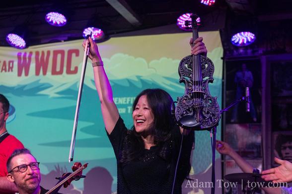 Emile K smiling with arms raised above her head, holding her ornate violin in one hand and her bow in the other. Ken Ferry is in the lower left of the photo, smiling at Emilie. Photo By Adam Tow.