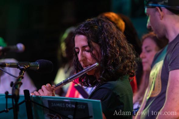 Sommer Panage playing flute, looking downward at sheet music. A number of band members stand behind her. Photo by Adam Tow.
