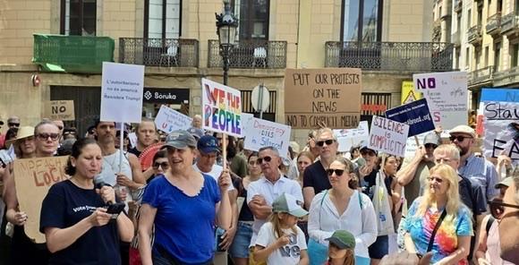 A group of people holding protest signs. They are standing in front of a light-colored stone building with ironwork balconies. The person at the front is speaking into a microphone. 