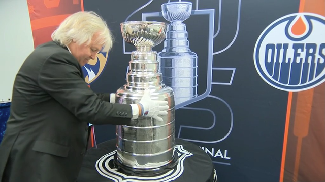 A man with shoulder-length white hair, wearing a black suit and white gloves, carefully places his hands on the Stanley Cup trophy. The iconic silver trophy sits on a black cloth-covered pedestal in front of a promotional backdrop for the NHL Stanley Cup Final. The backdrop features a large image of the trophy, the word “Final,” and team logos for the Edmonton Oilers and Florida Panthers. The scene captures a solemn, respectful moment as the trophy is being presented or prepared for display.