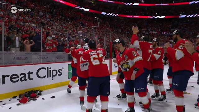 Florida Panthers players in red jerseys celebrate on the ice in front of a roaring home crowd. The players are hugging and raising their arms in joy, some tossing gear on the ice. Fans behind the glass wave and cheer, many wearing red. One player, Marchment (