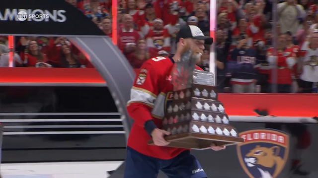 The same Florida Panthers player walks across the ice proudly holding the Conn Smythe Trophy. He beams with joy as fans in red jerseys cheer in the background. The arena glows with bright lights, and the “Florida Panthers” logo is visible on the boards. The player’s white cap reads “Champions,” marking the team’s triumphant moment as the crowd erupts with applause.