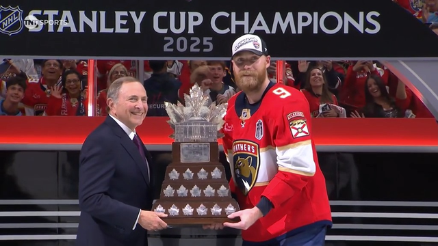 A Florida Panthers player wearing a red jersey and white championship cap stands beside NHL Commissioner Gary Bettman, both smiling while holding the Conn Smythe Trophy. A banner overhead reads “Stanley Cup Champions 2025.” The trophy is large, wooden-based, and topped with a silver leaf structure. Behind them, excited fans cheer and wave, dressed in team gear. It’s a formal moment honoring the playoffs’ MVP.