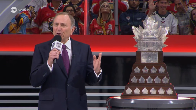 NHL Commissioner Gary Bettman speaks into a microphone while standing beside the Conn Smythe Trophy during the award presentation. He wears a dark suit and stands in front of a crowd of Florida Panthers fans, many in red jerseys and championship gear. The trophy is large and distinctive with silver maple leaves and a tall, leaf-shaped top. Bettman gestures as he makes the official announcement.