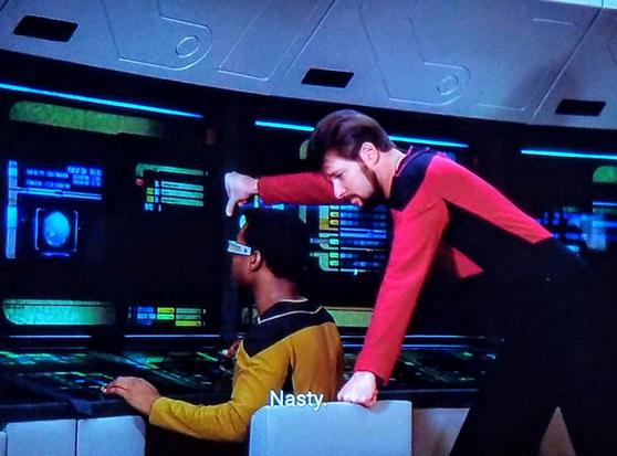 Star Trek The Next Generation scene. Geordi sits on the bridge at a workstation, consoles of computer input screens at his fingertips and along the wall. Riker, a bit close from a personal bubble standpoint, is standing just behind, with an arm resting on the wall above geordi's head and his other on the back of Geordi's chair. Closed caption reads, "Nasty."
