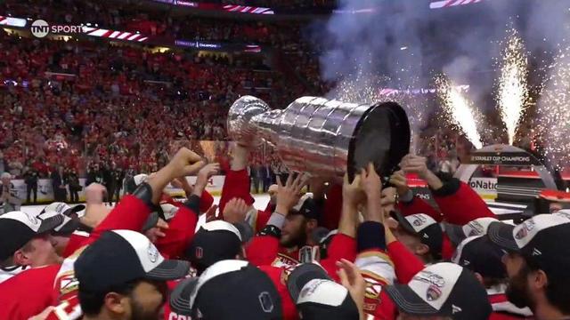 A close-up view of Florida Panthers players in red jerseys and black-and-white championship caps crowding together, all reaching up to touch the Stanley Cup. The trophy is suspended above their heads as a symbol of their triumph. The background shows thousands of fans in a packed arena celebrating. The image captures unity, emotion, and the joy of winning the NHL’s most prestigious prize.