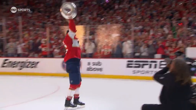 A Florida Panthers player skates on the ice, raising the Stanley Cup high above his head in celebration. He wears a red jersey and dark hockey pants, with fans packed in the background cheering wildly. Behind him, golden sparks burst into the air, and a professional photographer kneels nearby capturing the moment. The arena is filled with energy and red team colors after their 2025 Stanley Cup victory.