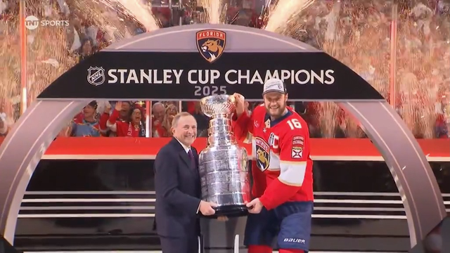 Under an arch that reads “Stanley Cup Champions 2025” with the Florida Panthers logo, NHL Commissioner Gary Bettman stands beside team captain Aleksander Barkov. Both smile as they hold the Stanley Cup trophy together. Barkov wears a red jersey and cap while golden fireworks erupt behind them. Fans in the background cheer ecstatically during this official presentation moment.